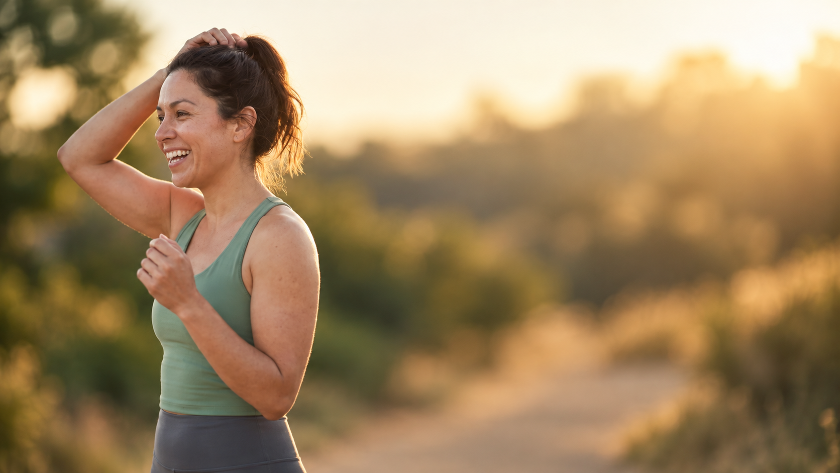 A woman tying her hair back on a sunlit trail at golden hour
